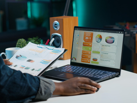 Woman sitting at home office desk, looking at business documents. African american freelancer in living room using notebook while reading financial charts on clipboard, camera A